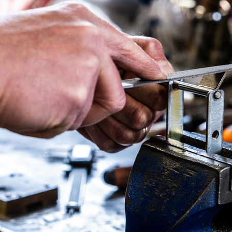Close-up of hands using a metal file to smooth the edge of a small metal bracket held in a bench vise, with various tools blurred in the background.