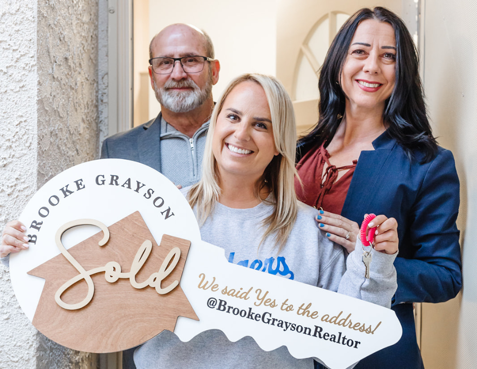 A young single woman holds an oversized key sign that says 'We said yes to the address,' with real estate agents Brooke and Jim standing proudly behind her, in front of the front door to her new home. Celebrating the journey of single women buying homes.
