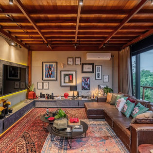 Sala de TV de casa de campo com assoalho e forro em madeira — o forro é o próprio piso do mezanino do pavimento superior —, móveis em couro e parede em painel de concreto aparente, em uma mistura de estilos que resulta em uma obra sofisticada e contemporânea