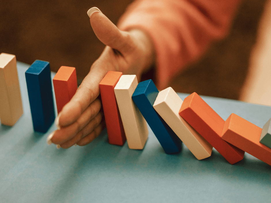 A hand reaching towards a line of colorful wooden blocks, preventing them from toppling over. The blocks are in red, blue, and cream colors.