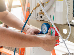 Technician adjusts blue and red pressure gauges on an outdoor HVAC unit, with hoses and wiring visible in bright sunlight.