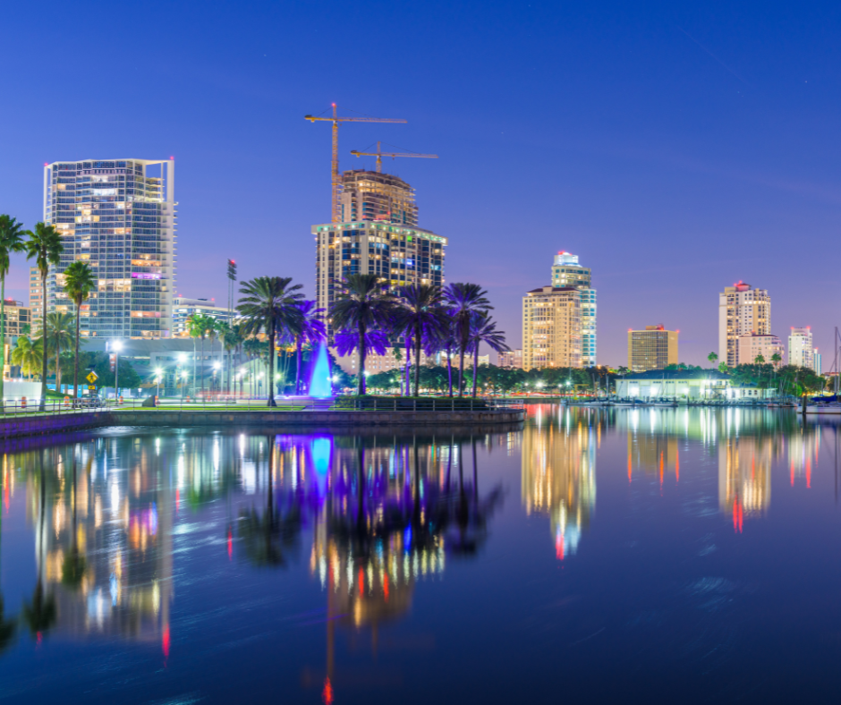 Night city skyline with palm trees, cranes, and glowing high-rises reflected in calm water under a blue dusk sky.