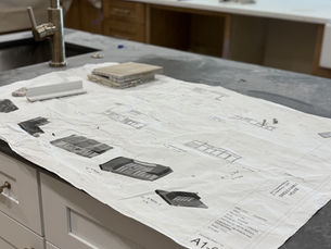 Blueprints on a kitchen counter display architectural designs. Tiles and hardware lie nearby. The setting is modern and neutral-toned. The picture and plans were made by HB Design and Engineering in Florida and it represents an Architectural drafter.