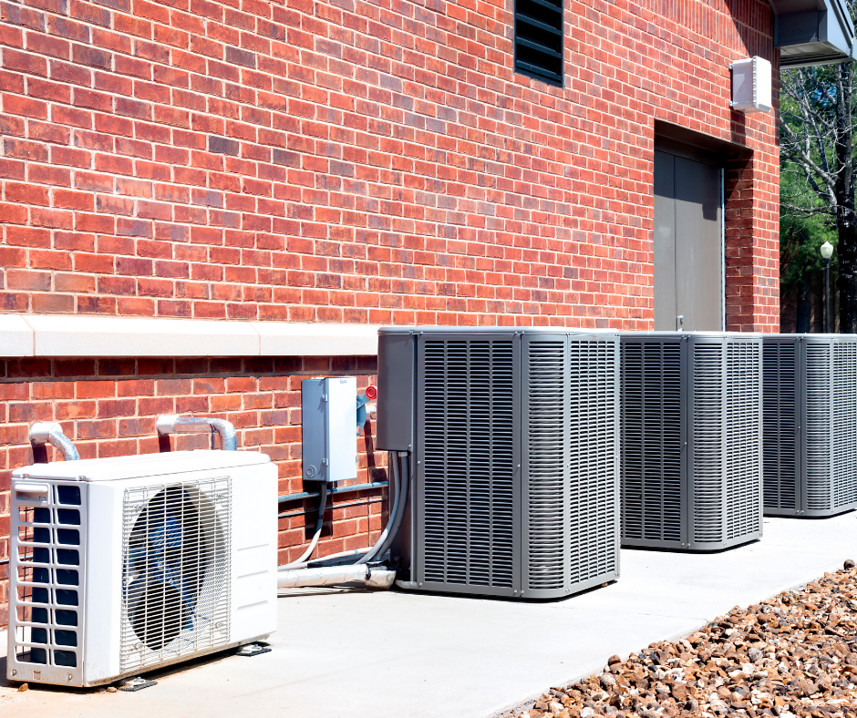 Row of outdoor HVAC units beside a red brick building on a concrete pad, with rocks and trees in the background.