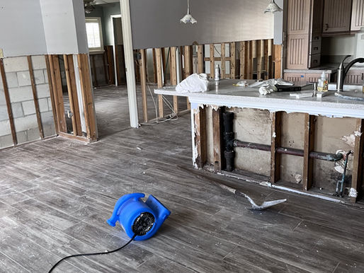 Partially demolished kitchen with exposed walls, a blue fan on wood floor, and minimal furnishings. Bright, under renovation setting. The image was taken by Built with Love in St. Petersburg to represent general contractor kitchen remodel