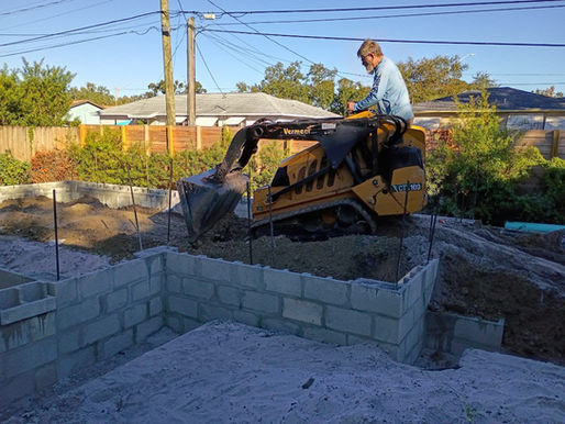 Man operates a yellow mini excavator in a backyard construction site. Blocks, sand, and greenery in the background under a clear blue sky. The picture was taken in St. Petersburg, Florida by Built with Love, exterior remodeling contractors.