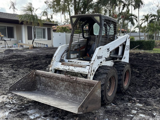 A small white Bobcat loader on a muddy ground near a house with windows, palm trees in the background. Overcast sky. No people visible. The image is used in a blog that describes general contractor vs builder.