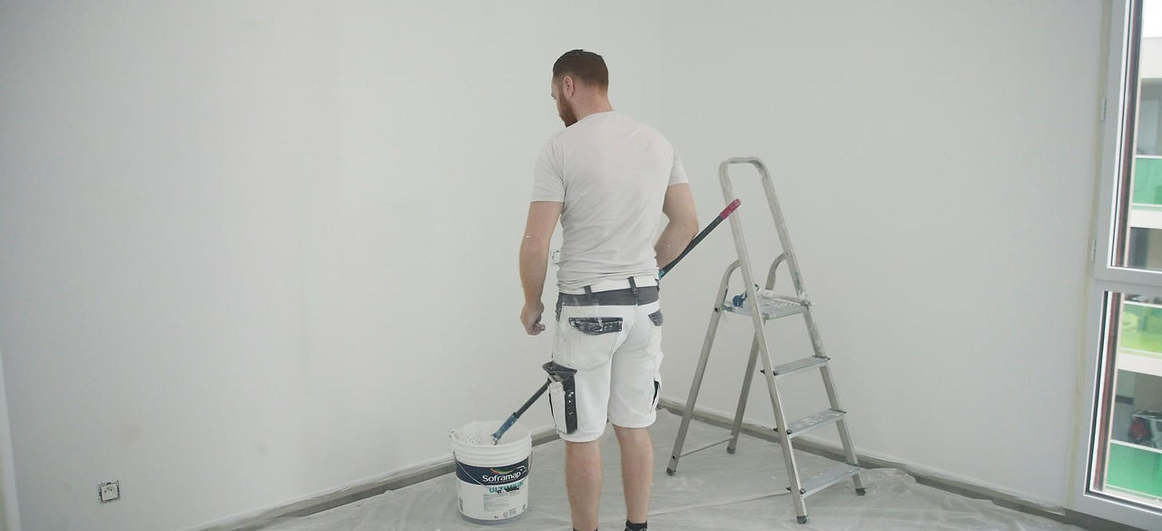 Cette image un artisan peintre en batiment de dos, face a un mur. il tiesn dans sa main une perche telescopiqueavce un rouleau de peinture en son extremité. il est entrain de tremper le rouleau dans un sceau de peinture. Le peintre en batiment est habillé avec un t-shirt blanc et un short blanc.