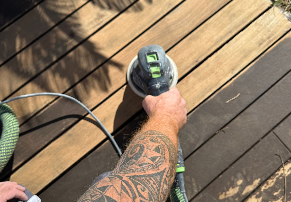 Ovanifrån av en tatuerad hantverkare som använder en elektrisk slipmaskin för att utföra slip- och skalningsarbete på ett trädäck. Bilden visar en markant kontrast mellan den del av träet som redan slipats, vilket avslöjar en ljus och ren yta, och den ännu inte behandlade delen, mörkare och sliten.