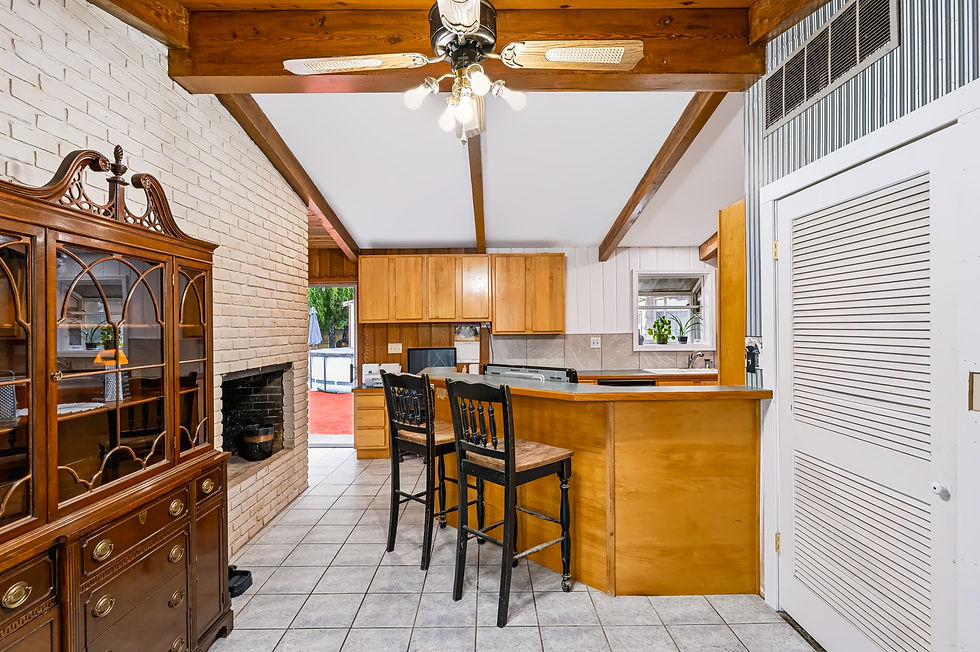 Open kitchen and dining area with exposed wood beam ceiling, brick fireplace, wood cabinetry, breakfast bar seating, and tile floors.
