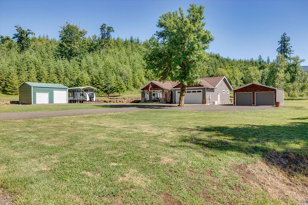 Home at 6620 Gold Creek Road, Willamina, OR, with brown shingle and stone exterior, attached two-car garage, covered front porch, and gravel driveway, surrounded by green lawn and forested hillside, with a green metal shop building, RV parked under a carport, and a second detached garage on the property.