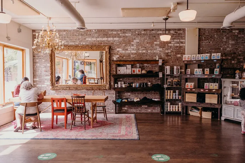 Cozy cafe interior with people at a wooden table, shelves of products, chandelier, and an OPEN sign on the mirror.