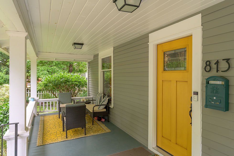 Front porch with yellow door marked 813, teal mailbox, wicker chairs on a yellow rug, and green trees beyond.
