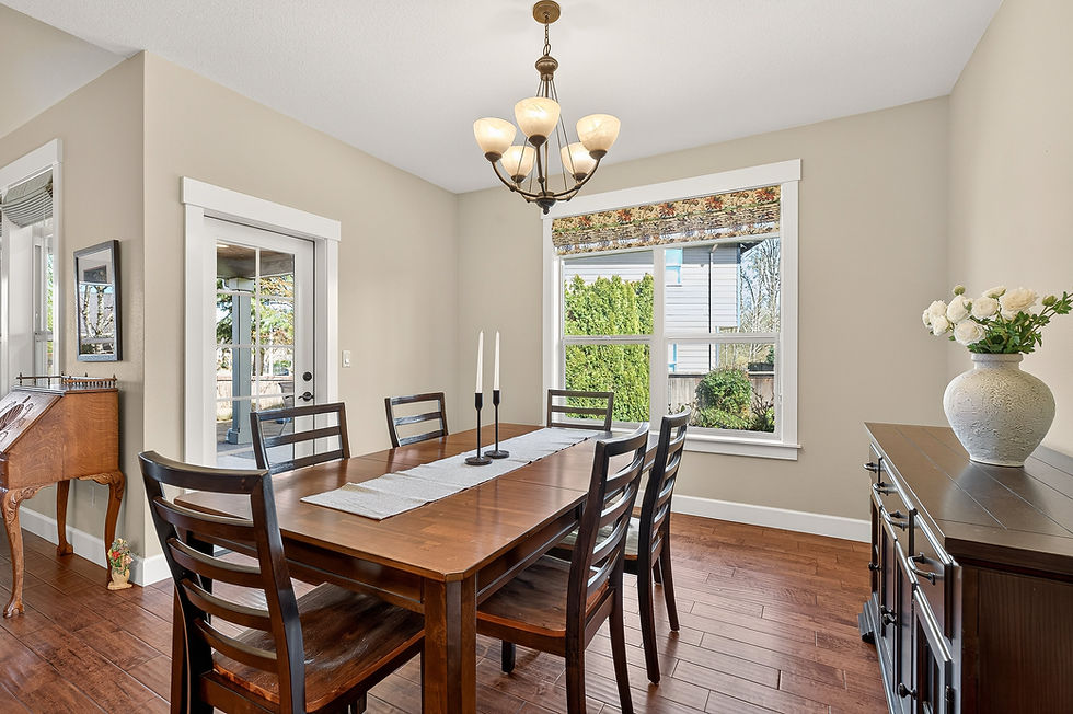 Dining room with a wooden table, six chairs, two white candles, and a chandelier. Large window with floral shade. Vase with roses on cabinet.