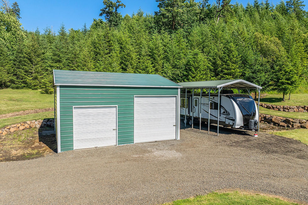 Green metal garage with white doors shelters a Reflection RV under a carport, on gravel with forested hills behind.