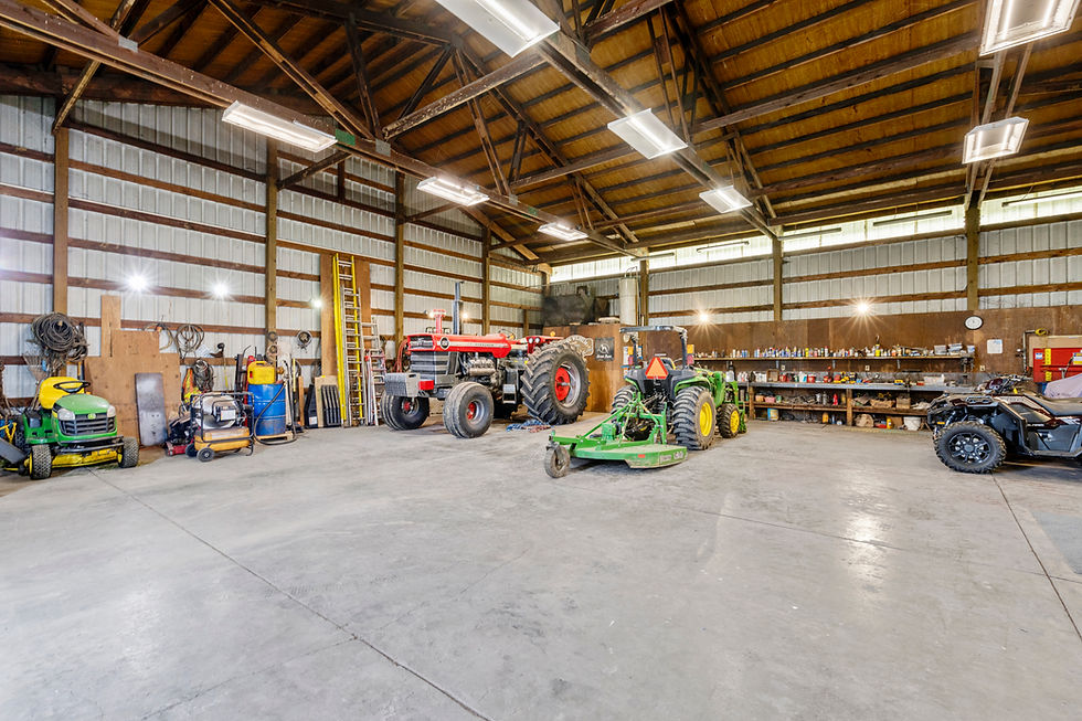 Spacious interior of a large metal-sided agricultural shop with exposed wood truss ceiling and fluorescent lighting, featuring a vintage red Massey Ferguson tractor, a green John Deere tractor with attached mower implement, a John Deere riding lawn mower, an ATV/side-by-side, and a long workbench with tools and supplies along the right wall, all on a polished concrete floor.