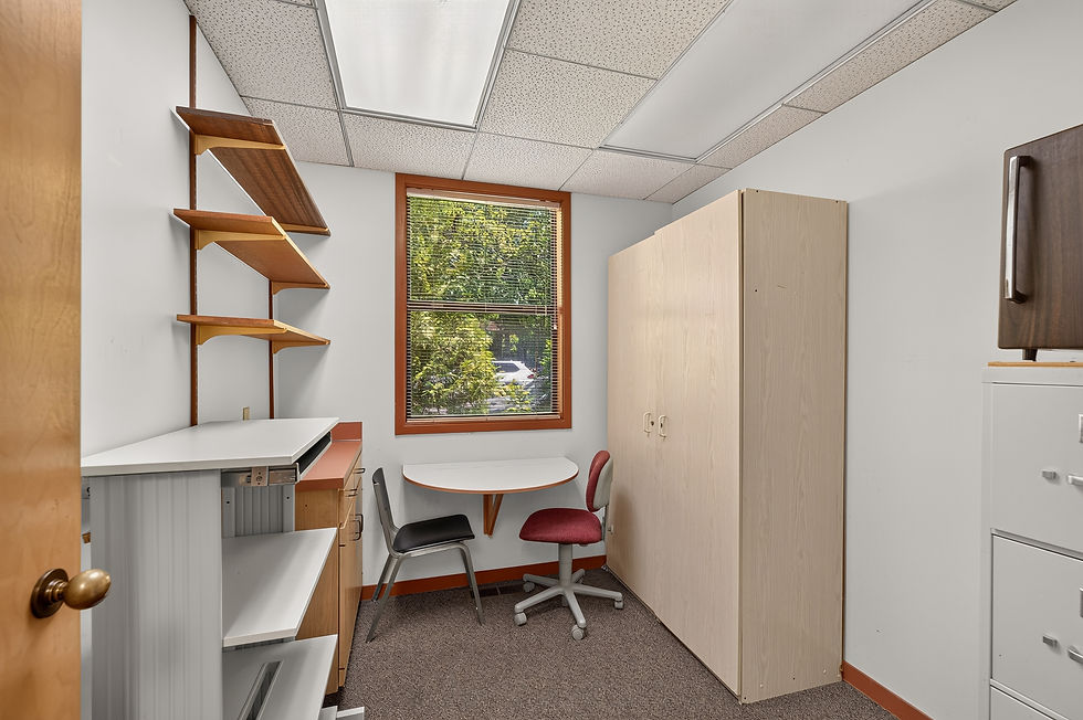 Empty small office/exam room with two chairs, desk, shelves, cabinets, and a window showing trees outside.