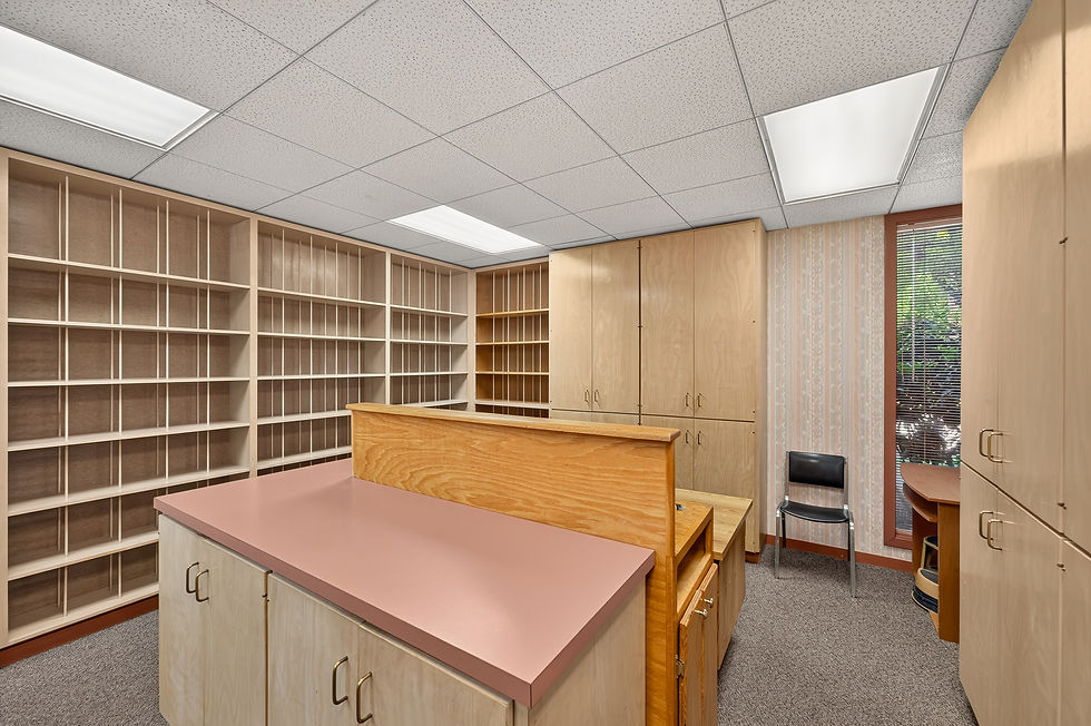 Empty office reception with wood shelves, pink counter, cabinets, and a chair by a window with blinds and patterned wallpaper.