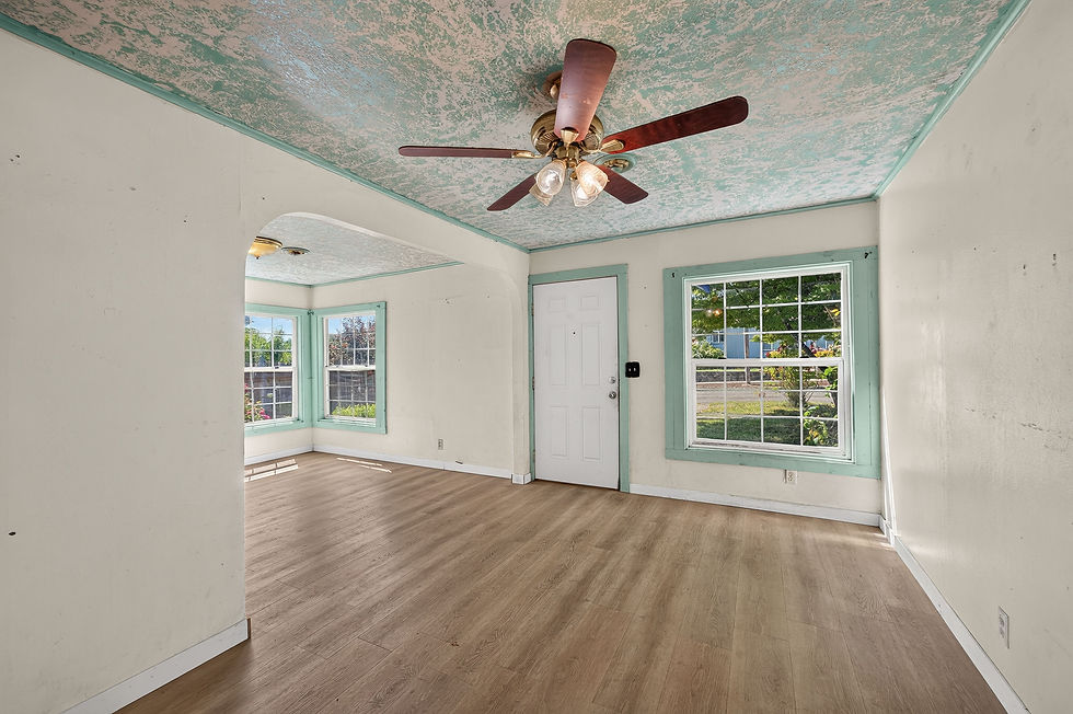 Empty living room with wood floor, mint-trimmed windows and door, and a ceiling fan under a mottled teal ceiling, bright daylight.