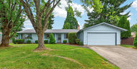Single-level ranch home at 127 NW 19th St McMinnville Oregon, light blue siding, attached two-car garage, mature shade trees, and lush green front lawn on a sunny day.