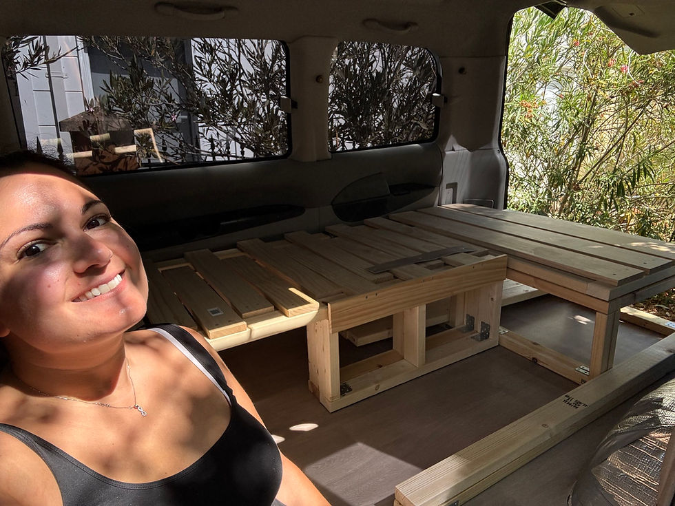 Smiling person in a van with a wooden DIY bed frame. Sunlit interior, trees visible through windows, relaxed and content mood.