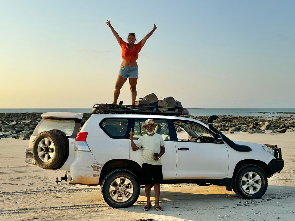 Woman stands on a white SUV roof, arms raised, on a rocky beach in the Dampier Peninsula. Another person stands beside the car, smiling. Clear sky, calm sea.