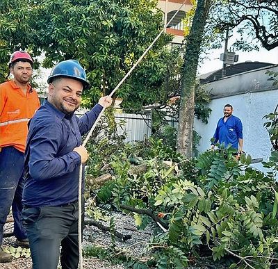 "Homens realizando o corte e remoção de uma árvore em ambiente urbano. No primeiro plano, um trabalhador com capacete azul 