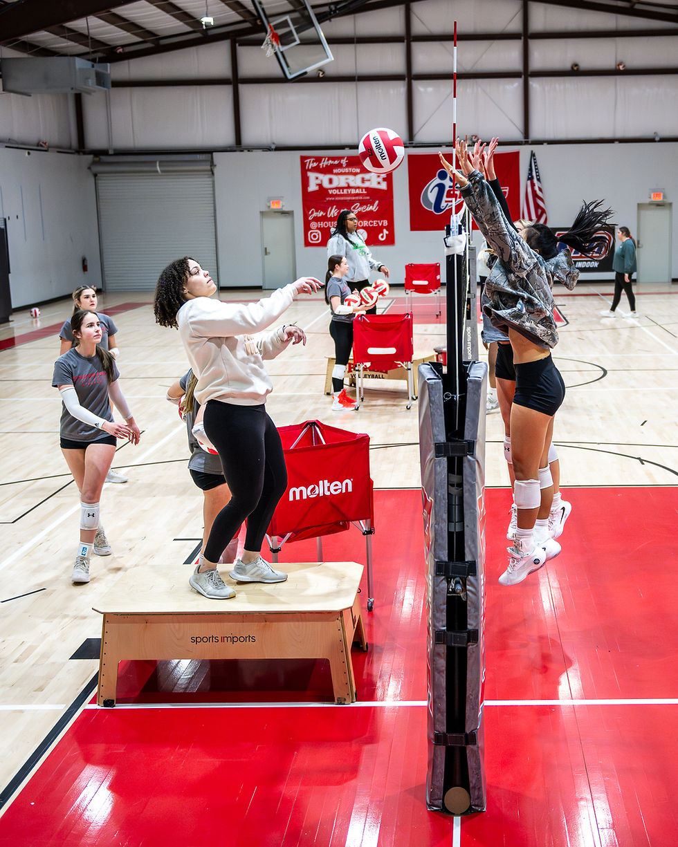 Two volleyball players jump at the net in a gym. One is on a sports platform. Red and white decor, with visible team logos in the back.