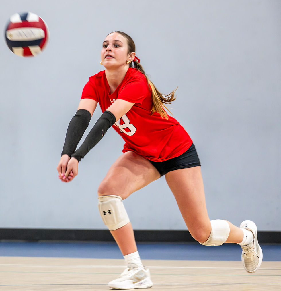 Volleyball player in red jersey and black shorts diving for a ball in a gym. Focused expression, knee pads, neutral background.