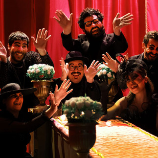 Grupo de pessoas comemorando e sorrindo ao redor de uma mesa decorada, com cortina vermelha de fundo, em momento de celebração e alegria