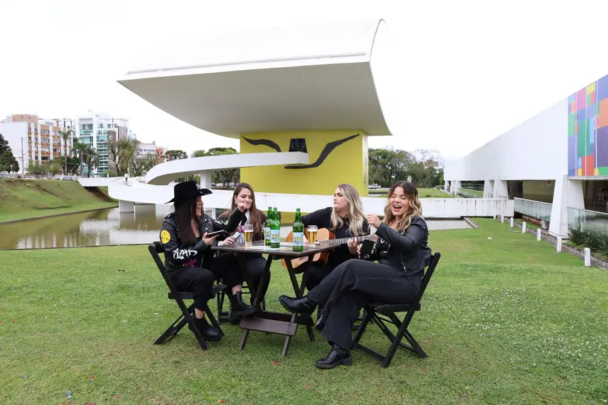 Grupo de quatro mulheres sorrindo e tocando violão em uma mesa ao ar livre no parque, com prédio moderno ao fundo, em uma tarde ensolarada.