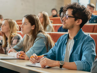 Students attentively listen in a lecture hall with red seats and notebooks. The setting is bright, and the mood is focused.