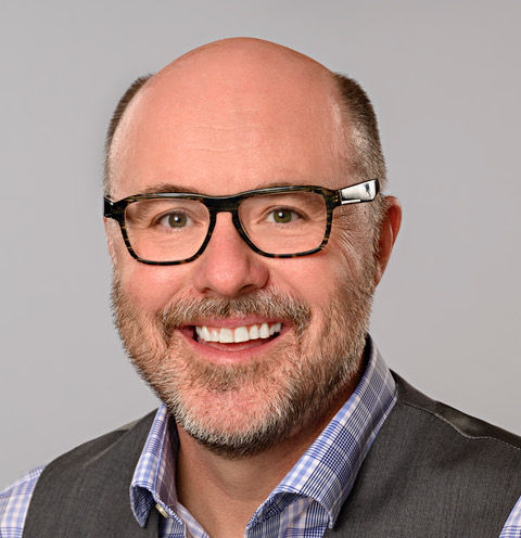 Professional headshot of Kevin, smiling and wearing glasses, a checkered shirt, and a gray vest against a light gray background.