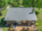 Aerial view of a brick house with a gray metal roof, wooden deck, and patio furniture. Lush green yard and trees in the background.