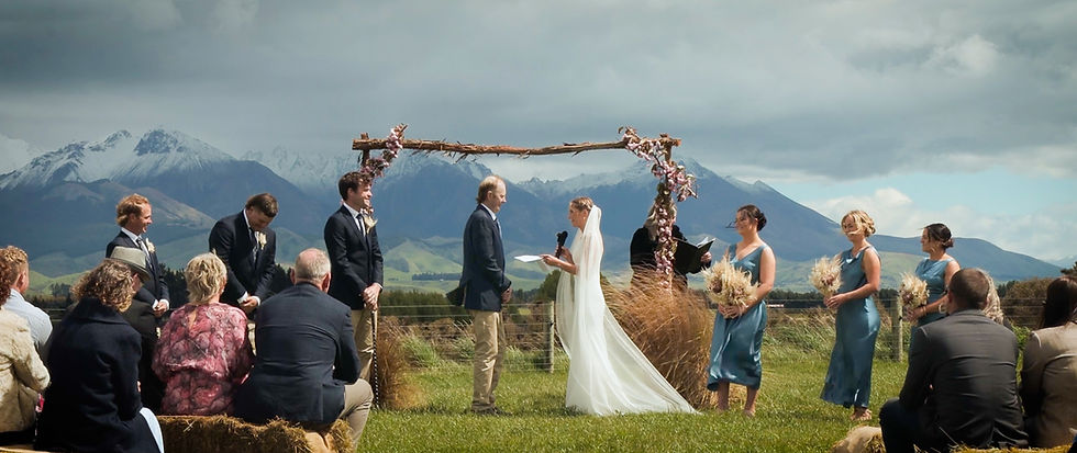 Outdoor wedding ceremony with a bride in white and groom under an arch. Guests seated on hay bales, mountains in the background.