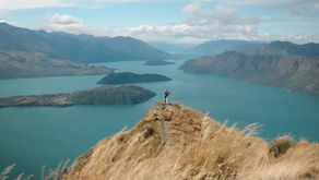 Couple embraces on a grassy cliff overlooking a turquoise lake and mountains, under a blue sky with scattered clouds. Majestic and serene at Roys Peak, Wanaka, New Zealand