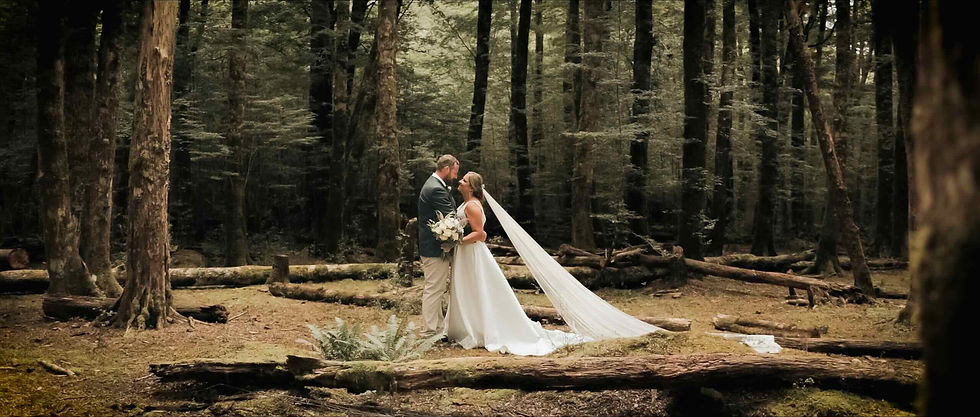 Bride and groom embracing in a forest. Bride holds white flowers, veil flowing. Sunlight filters through tall trees, creating a serene mood.
