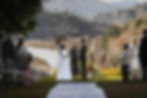 Bride and groom stand under a floral arch during an outdoor wedding ceremony with mountains in the background. Guests are seated, observing.