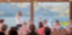 Man in white shirt gives a speech at a wedding, others raise hands joyfully. Mountain lake view; hanging lights, flowers add warmth.