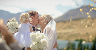 Cinematic wedding moment capturing the bride and groom’s first kiss outdoors with their child, surrounded by white flowers and golden petals against a mountain backdrop in New Zealand