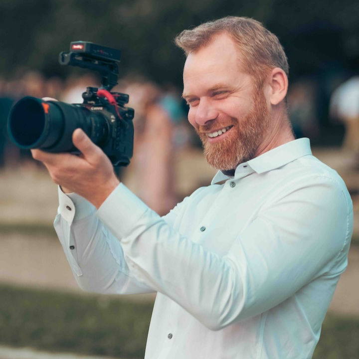 Smiling bearded man in a white shirt holding a professional camera outdoors, aiming it with a cheerful, focused expression