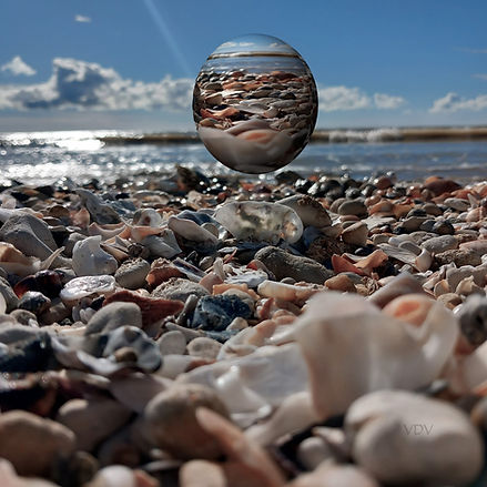 Une photographie d'une plage de galets et coquillages. Au centre se trouve une sphère qui reflète la plage.