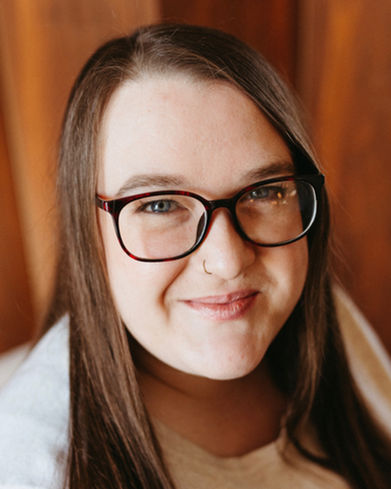 Jane is wearing a light coloured sweater and is smiling while looking directly at the camera. She is wearing large dark coloured glasses, has long brown hair, blue eyes, and a gold nose ring. The background is an out of focus, warm orange/brown wood.