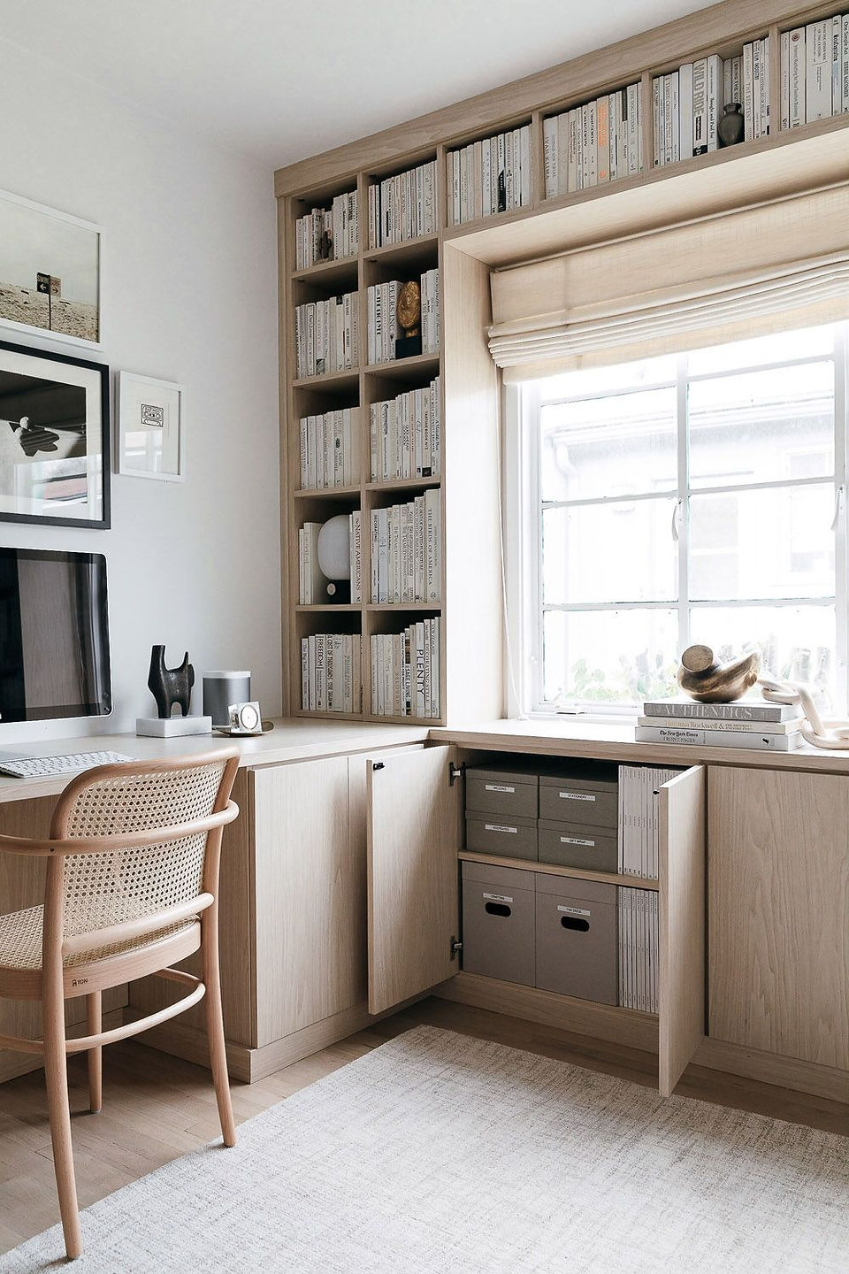 Bureau moderne en bois, avec une écran d'ordinateur, une chaise en cannage, une bibliothèque avec des livres et une fenêtre vers l'extérieur.