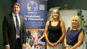 Mark Griffin MSP with Craighalbert’s CEO, Heather Gray and Chair, Mary Morgan, standing beside a colourful Craighalbert display featuring photographs of children and young people.