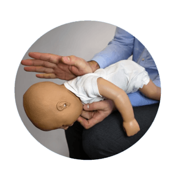 Eine Person führt an einer Babypuppe die Heimlich-Methode durch, um einen Fremdkörper aus den Atemwegen zu entfernen. Die Puppe liegt mit dem Gesicht nach unten auf dem Oberschenkel der Person, während die Hand der Person auf den Rücken der Puppe klopft. Diese Darstellung simuliert die Erste-Hilfe-Maßnahme bei Säuglings-Erstickungsnotfällen.