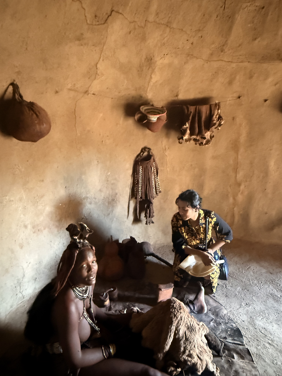 A Himba woman sits inside a traditional hut, engaging in conversation. The earthen walls display hanging cultural artifacts, reflecting the rich heritage of the Himba people.