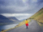 Sarmin in vibrant colors walks along a winding road, with dramatic mountains rising from the sea under a cloudy sky in the Faroe Islands.