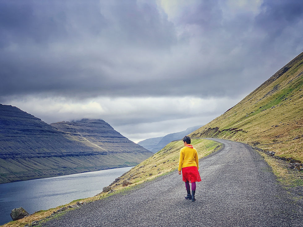 Sarmin in vibrant colors walks along a winding road, with dramatic mountains rising from the sea under a cloudy sky in the Faroe Islands.