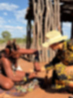 A Himba woman helps Sarmin try on handcrafted jewelry, beautifully showcasing the intricate beadwork and cultural artistry amidst a sunlit village backdrop.